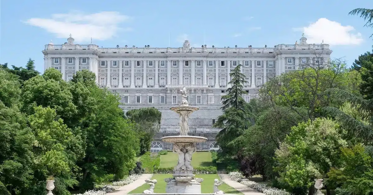 Madrid palais royal Le Palais Royal de Madrid, vu depuis ses jardins formels, avec une fontaine centrale ornée de statues et une végétation luxuriante autour.
