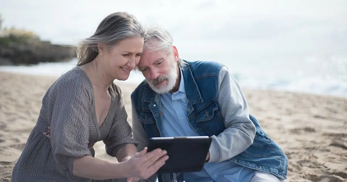 Couple de retraités assis sur une plage, consultant une tablette ensemble, illustrant une transition géographique douce vers l’Espagne sans rupture culturelle.